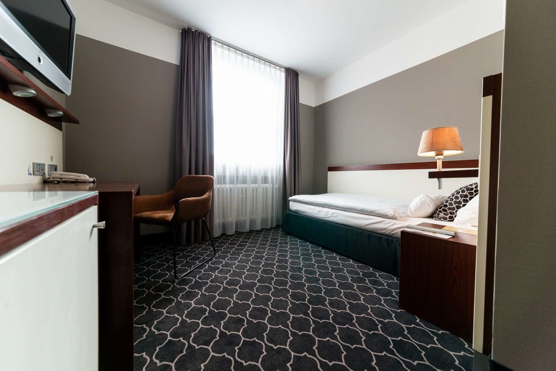 A hotel room with a bed, a desk with a telephone, a chair, a lamp, and a window with curtains.