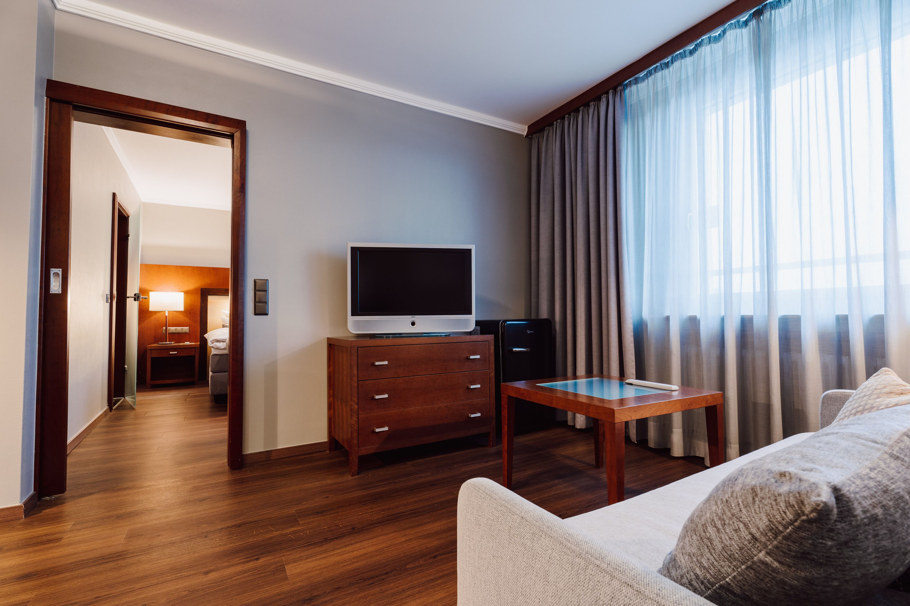 A hotel room with a wooden floor, a television on a wooden cabinet, a small table, a sofa, and a large window with curtains.