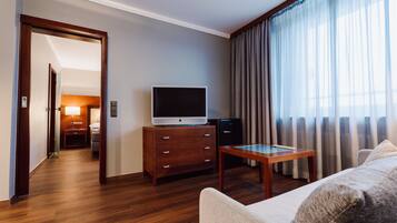 A hotel room with a wooden floor, a television on a wooden cabinet, a small table, a sofa, and a large window with curtains.