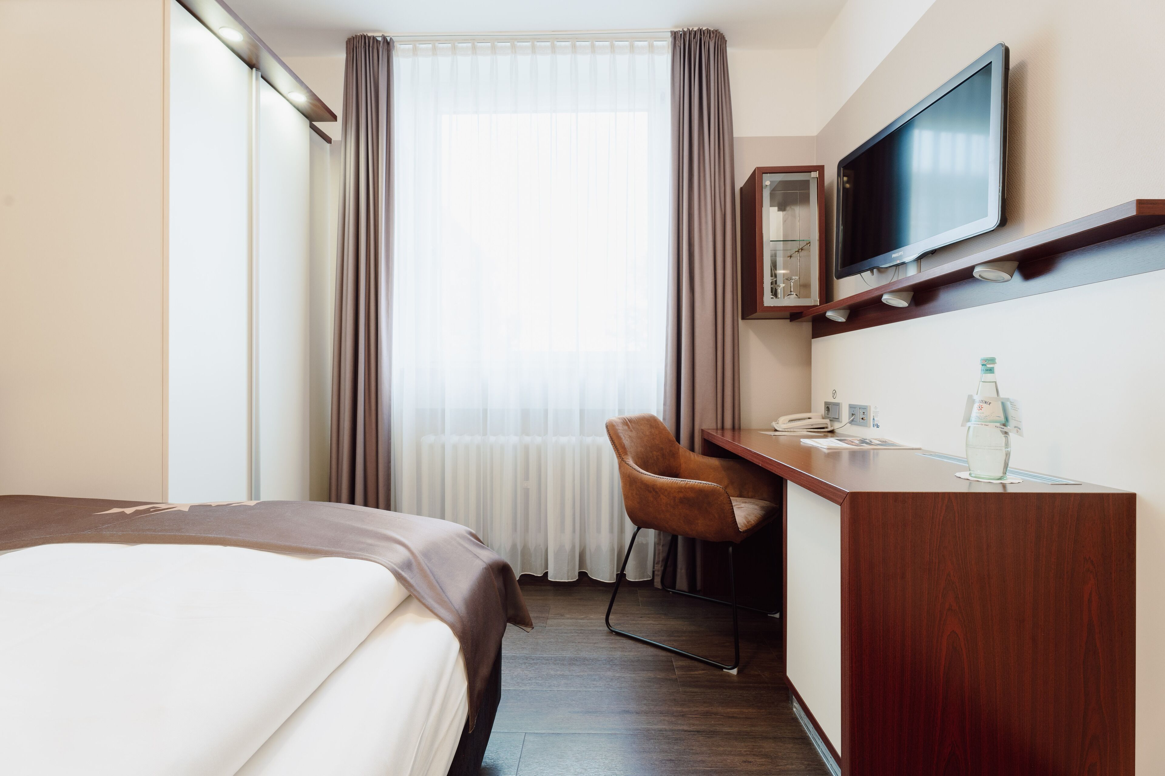 Habitación de hotel con cama, escritorio con silla, televisor montado en la pared y ventana con cortinas.
