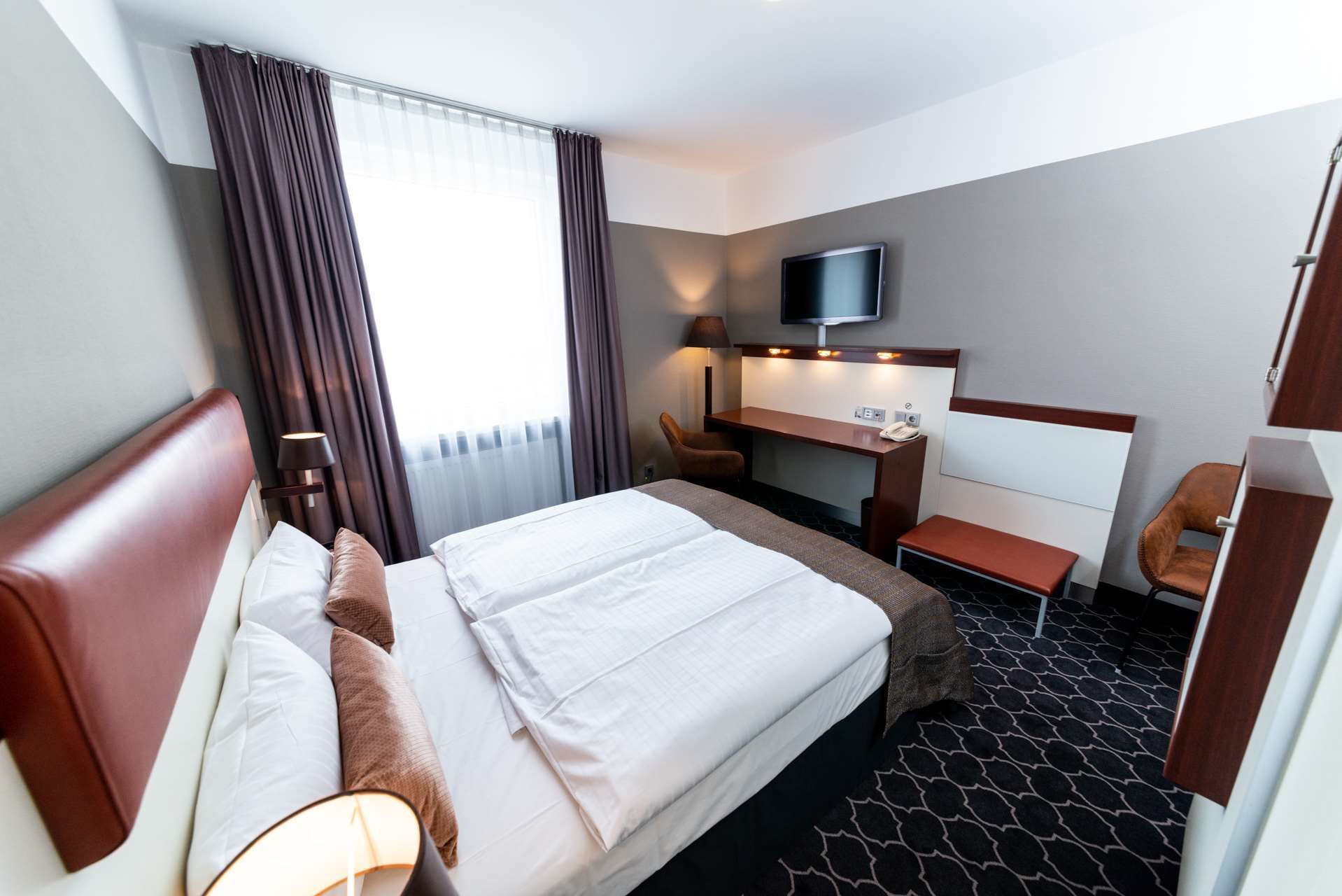 Une chambre d’hôtel avec un lit, un bureau, une chaise, une télévision et une fenêtre avec des rideaux.