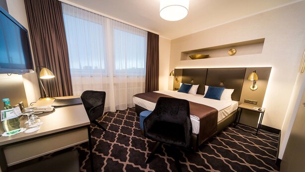 Habitación de hotel con cama, escritorio, silla, televisor y ventana con cortinas.