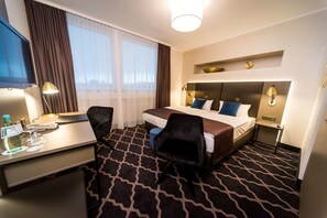 Habitación de hotel con cama, escritorio, silla, televisor y ventana con cortinas.
