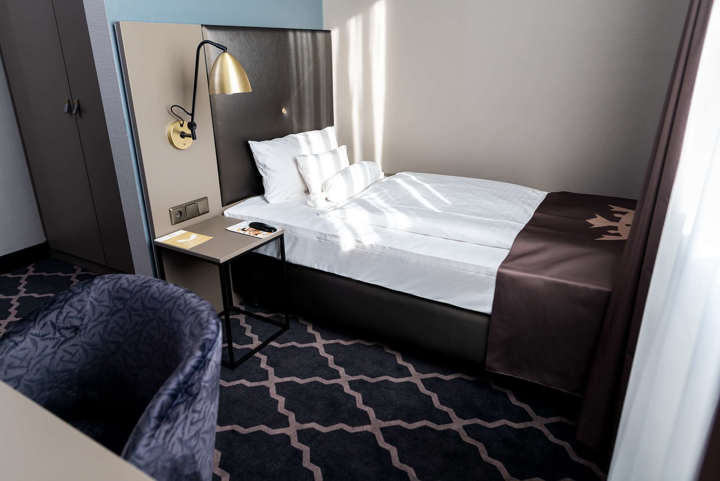 A hotel room with a bed, a bedside table, a lamp, a chair, and a patterned carpet.