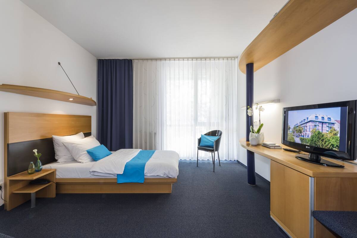 Een moderne hotelkamer met een bed, een bureau met een televisie, een stoel en een raam met gordijnen.