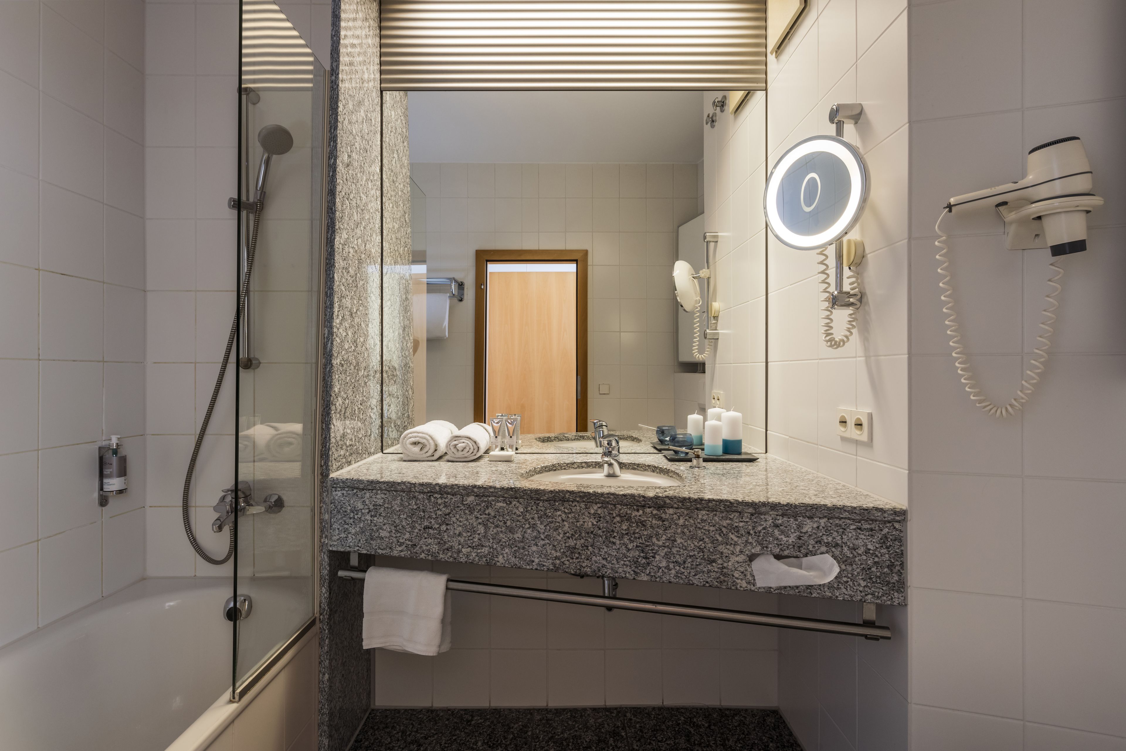 A bathroom with a glass-enclosed shower, a granite countertop sink, a mirror with a light, and a hairdryer mounted on the wall.