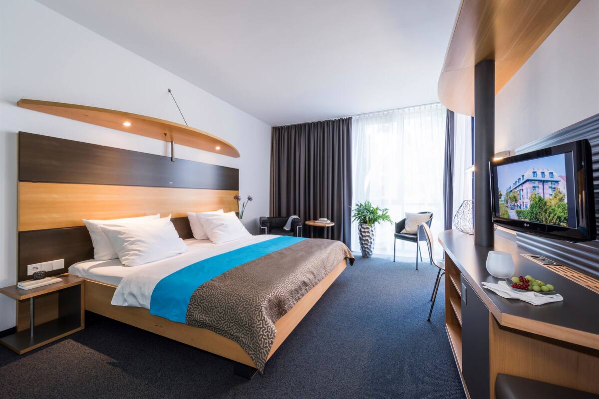 Een moderne hotelkamer met een groot bed, een bureau met een televisie, een klein tafeltje en uitzicht op de stad door grote ramen.