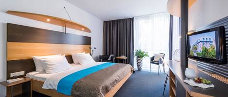 Habitación de hotel moderna con una cama grande, un escritorio con televisor, una mesita y vistas a la ciudad a través de amplios ventanales.