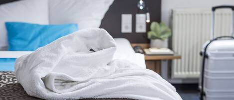A neatly made bed with a white towel, a blue pillow, and a patterned pillow. There is a white suitcase and a radiator in the background.