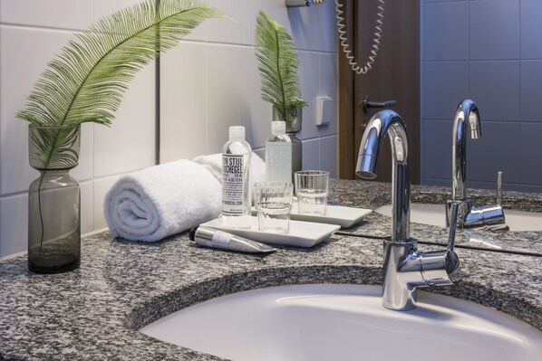 A bathroom with a granite countertop, a white sink, two chrome faucets, a towel, a glass vase with a plant, and a bottle of lotion. - SEEhotel Friedrichshafen (Friedrichshafen)