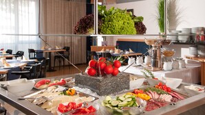 Un bufé con diversos alimentos, entre ellos frutas, verduras y carnes, en un comedor bien iluminado con mesas y sillas.