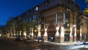 Un hotel con un diseño arquitectónico moderno, que destaca un letrero grande que dice 'See Hotel', un estacionamiento y vista a la calle por la noche.