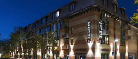 A hotel with a modern architectural design, featuring a prominent 'See Hotel' sign, a parking area, and a street view at night.