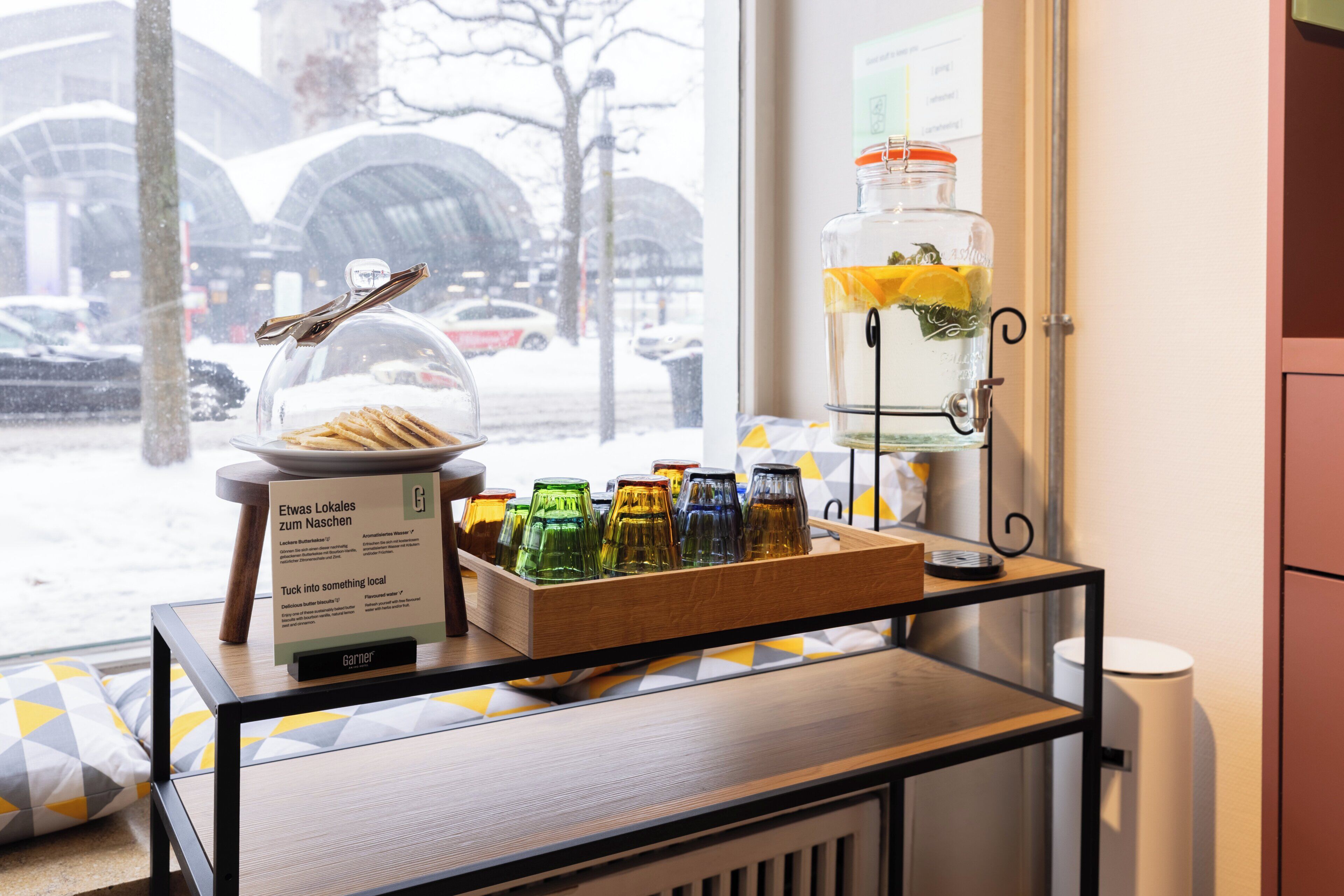 A breakfast buffet with a glass jar of lemon slices, a sign with local information, and a window view of parked cars and a tree.