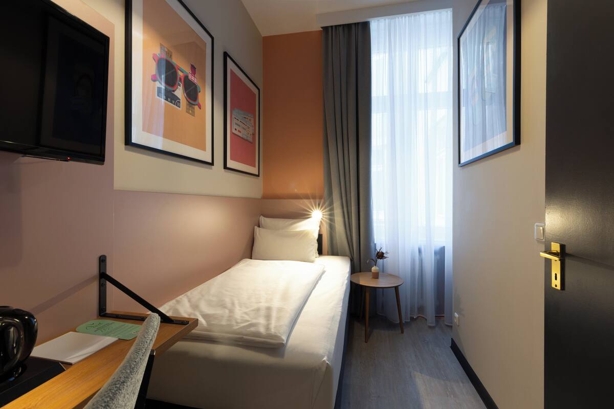 Ein Hotelzimmer mit einem Bett, einem Schreibtisch mit Stuhl, einem kleinen Tisch mit Vase und einem Fenster mit Vorhängen.