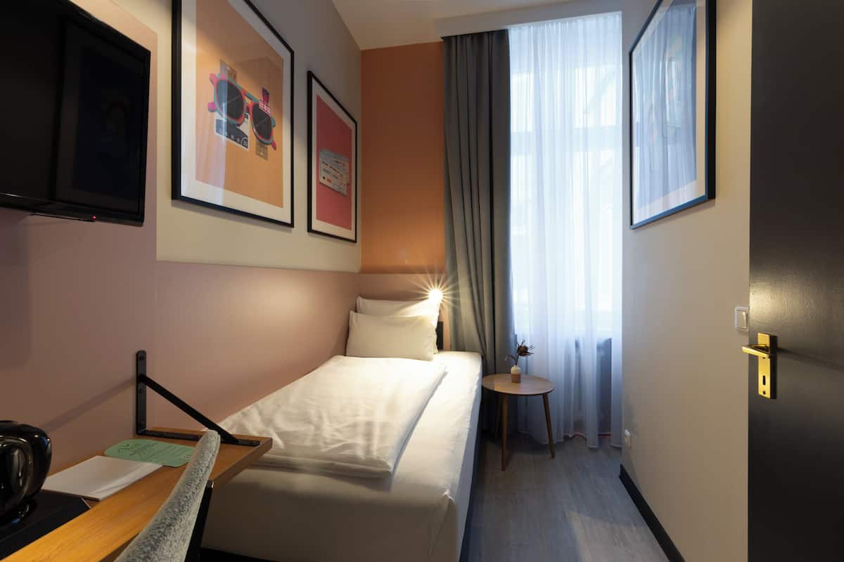 Et hotelværelse med en seng, et skrivebord med stol, et lille bord med vase og et vindue med gardiner.