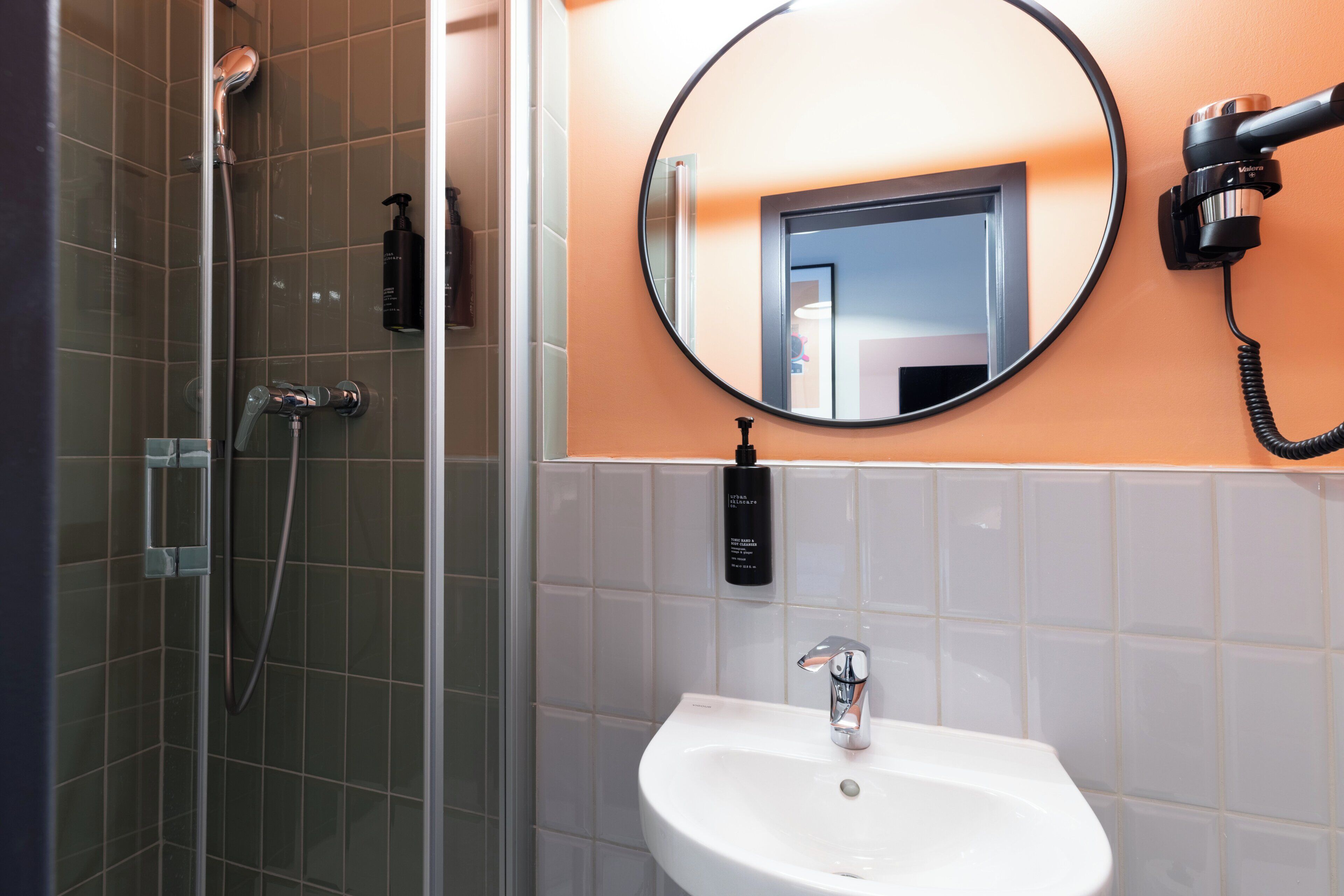 A bathroom with a glass-enclosed shower, a white sink, a round mirror, and a handheld hair dryer mounted on the wall.