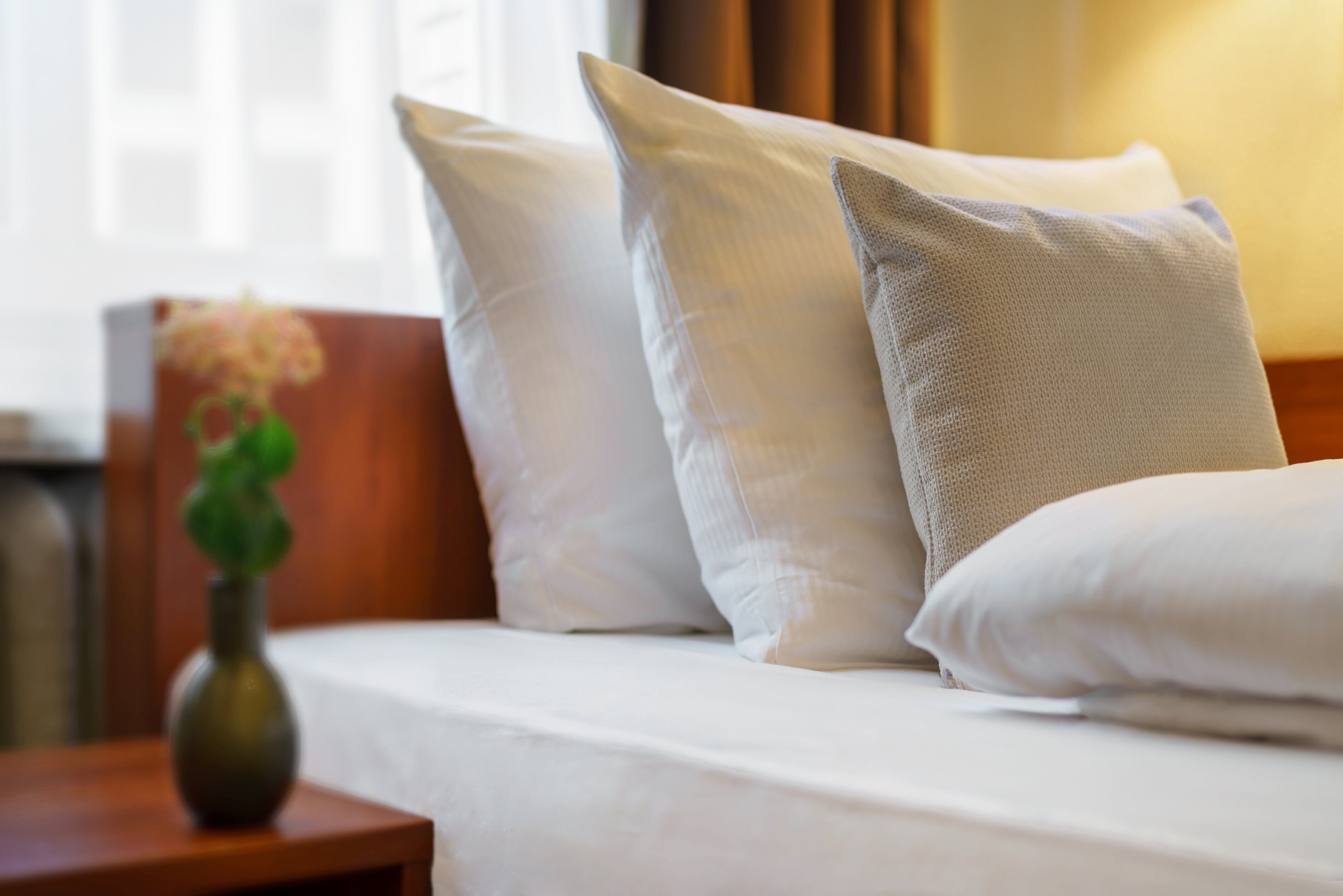 A bed with white linens and pillows, a wooden nightstand with a vase of flowers, and a window with curtains.