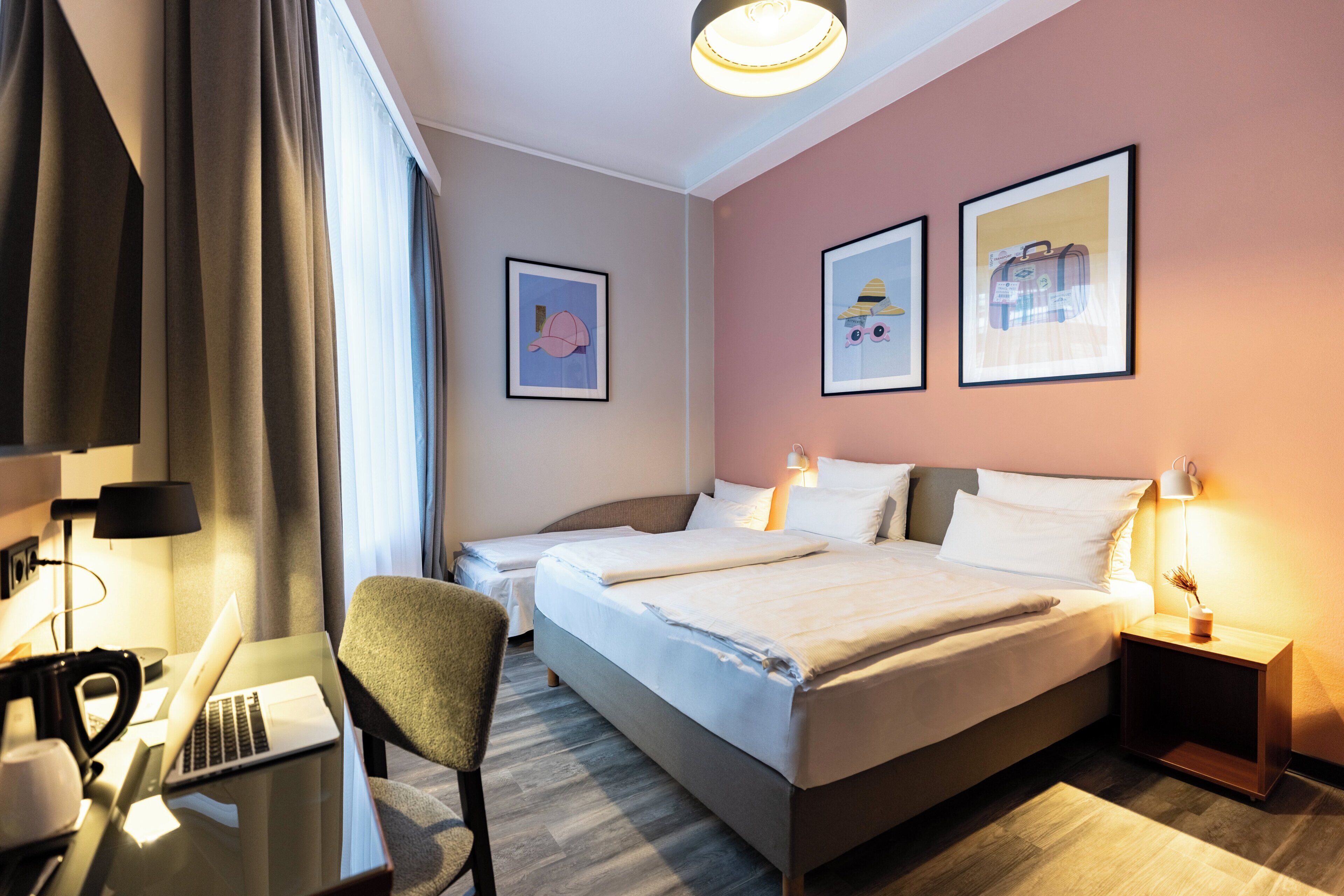 A hotel room with a bed, a desk with a laptop, a chair, bedside tables with lamps, and framed pictures on the wall.