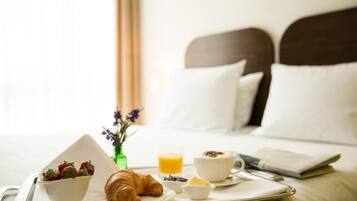 A breakfast tray with croissants, strawberries, a bowl of cereal, a glass of orange juice, and a croissant on a bed with pillows and a vase of flowers.