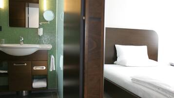 Habitación de hotel con cabecera de madera, cama blanca, baño con lavamanos, espejo y paredes de azulejos verdes.