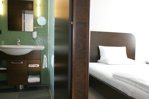 Habitación de hotel con cabecera de madera, cama blanca, baño con lavamanos, espejo y paredes de azulejos verdes.