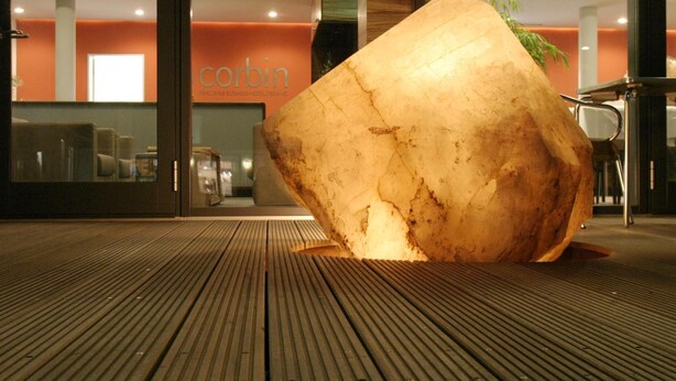 A large, illuminated sculpture in an outdoor area with a wooden floor and a building with a sign reading "Corbin" in the background.