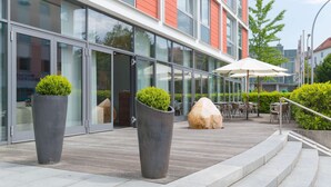A modern building with large glass windows, outdoor seating area with tables and chairs, and two large planters with greenery.