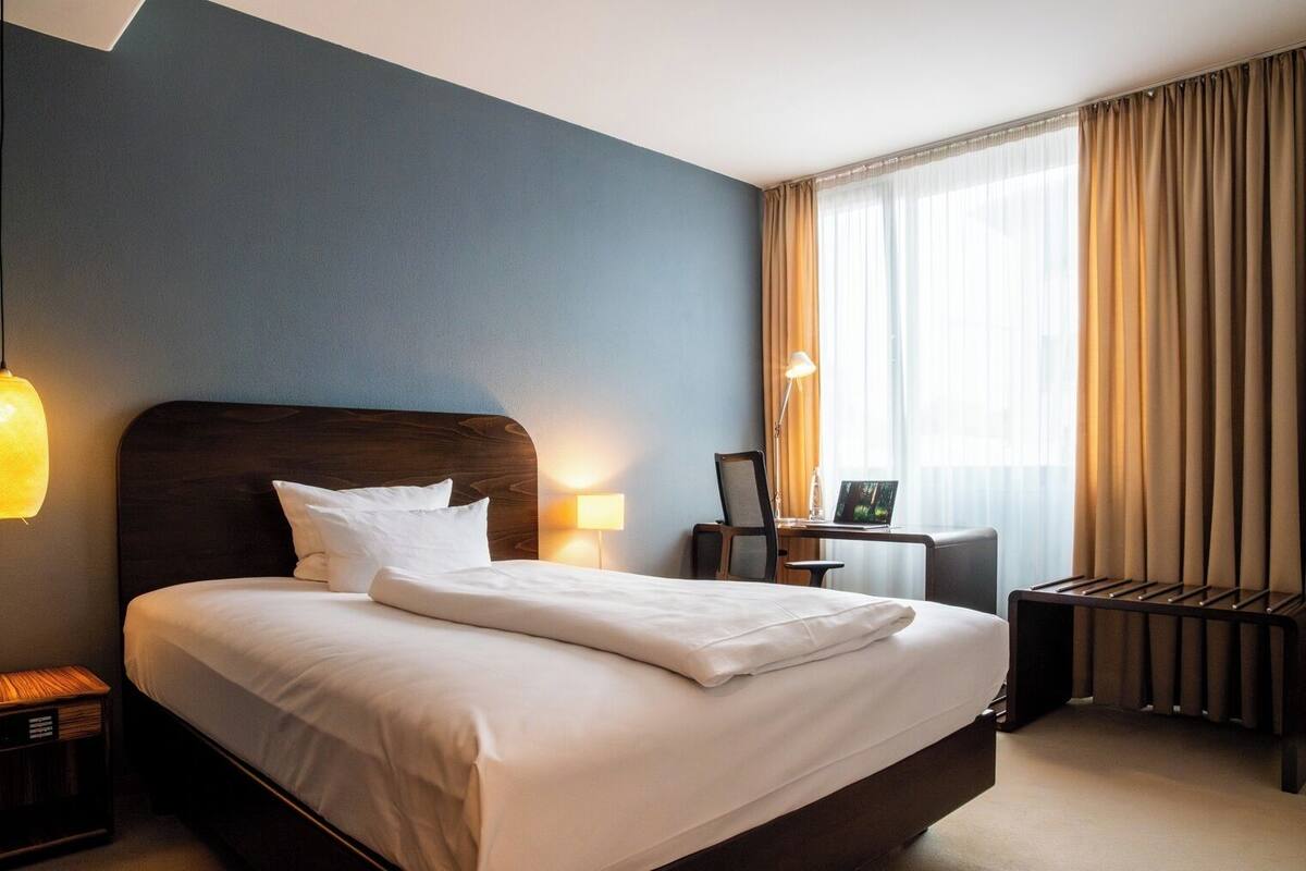 A hotel room with a large bed, a desk with a chair, a lamp, and a view of the outdoors through the window.