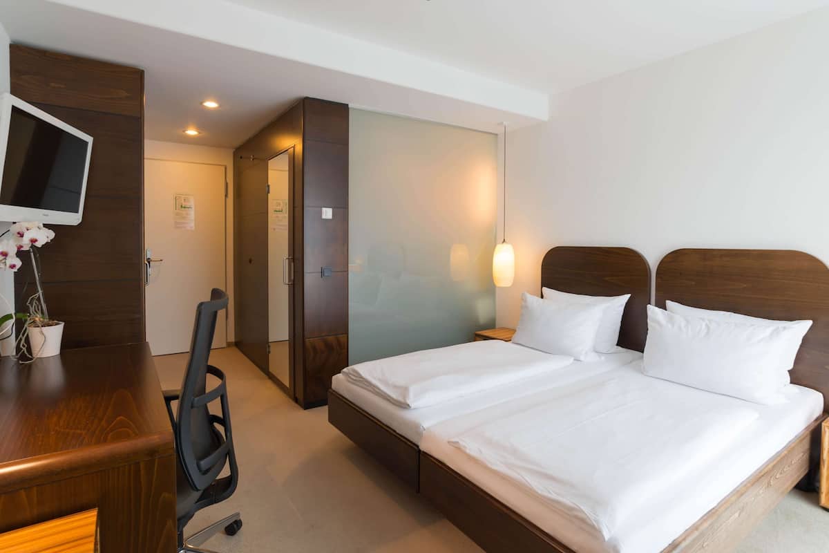 A hotel room with a bed, a desk, a chair, a TV, and a wall-mounted light.