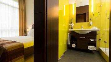 A hotel room with a bed, a bedside table with a lamp, a bathroom with a sink, mirror, and towel rack.