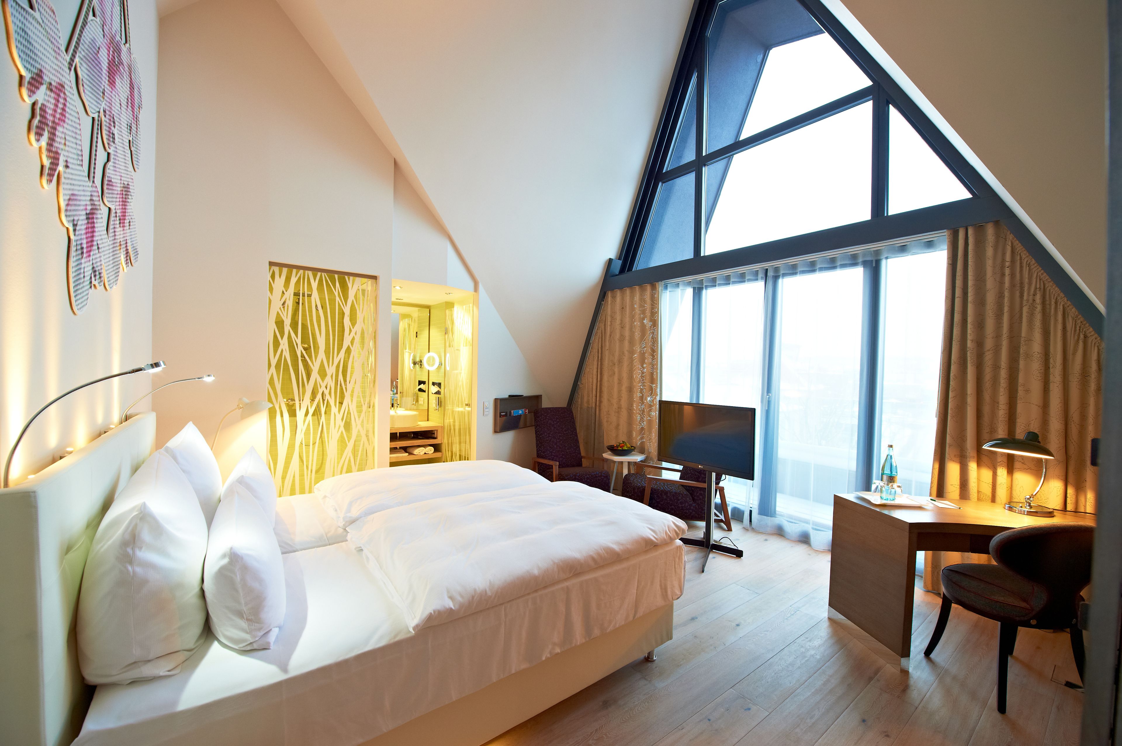 A modern hotel room with a large bed, a desk, a chair, and a large window with curtains.