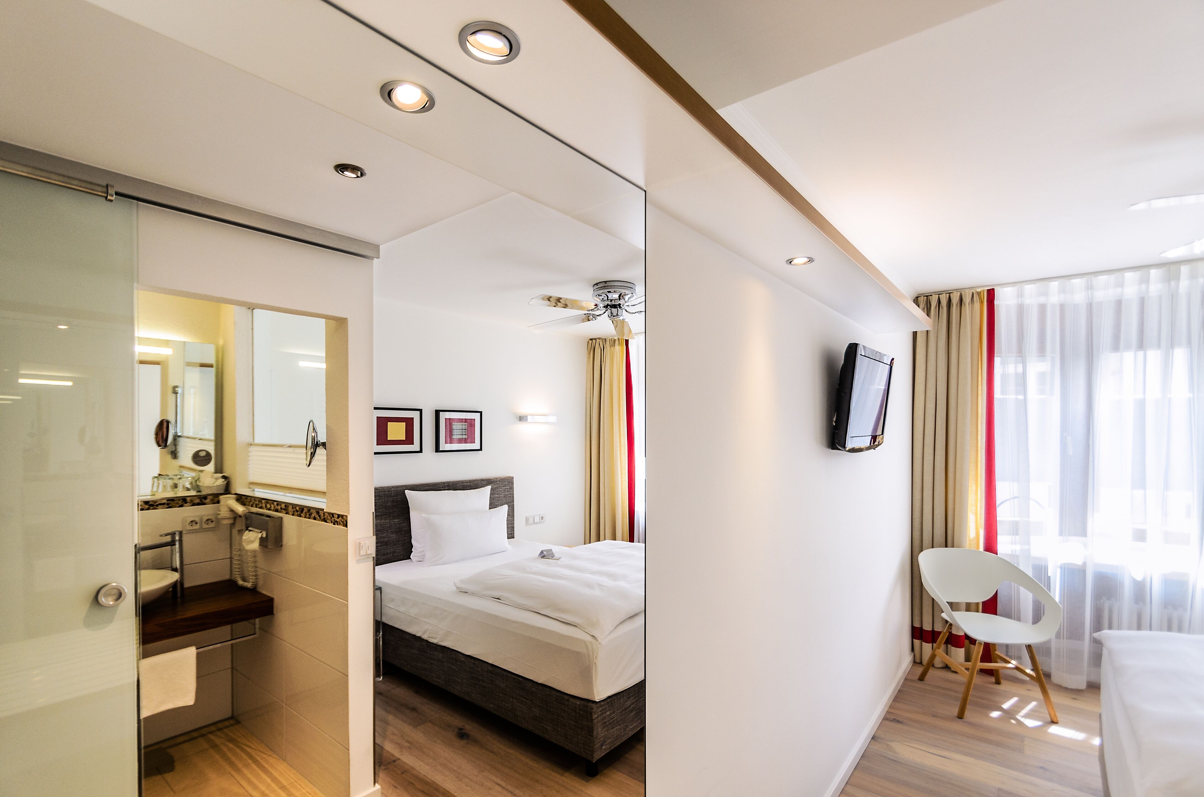 A modern hotel room with a bed, a bathroom, a TV, and a window with curtains.