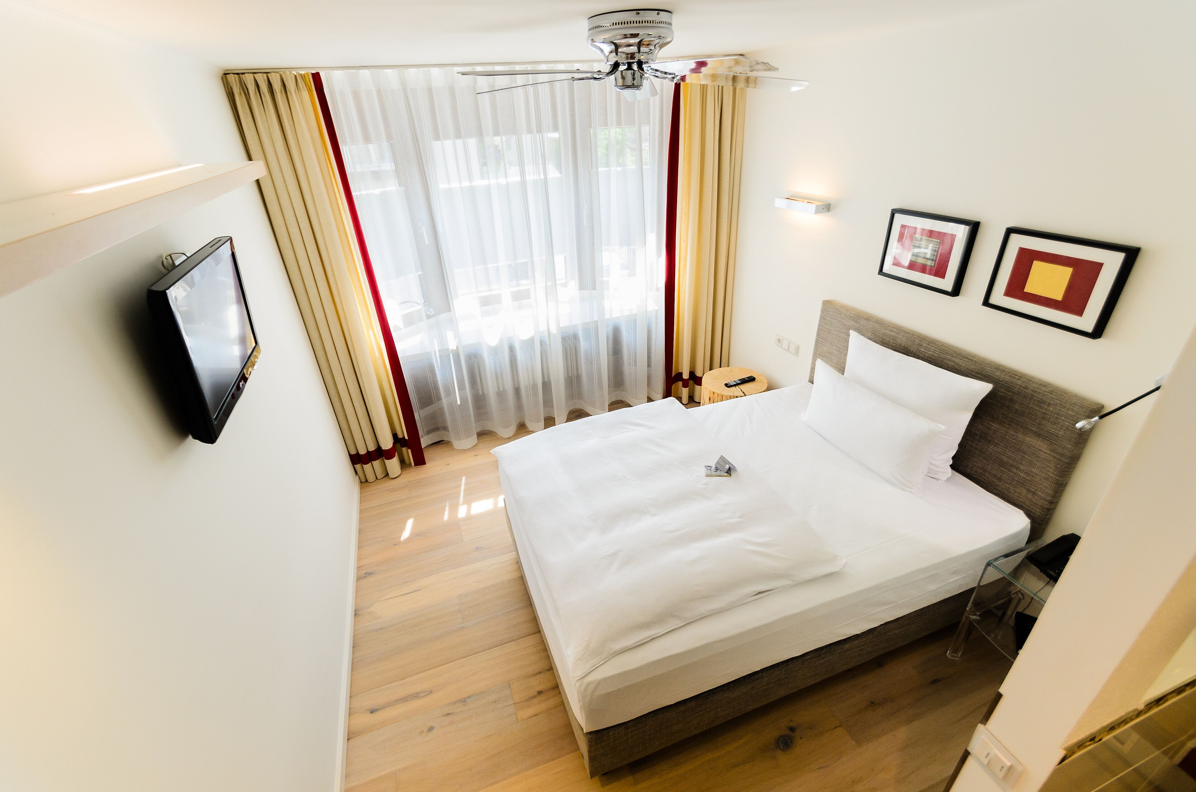 Ein Hotelzimmer mit Bett, Vorhängen, Deckenventilator, Fernseher und zwei gerahmten Bildern an der Wand.