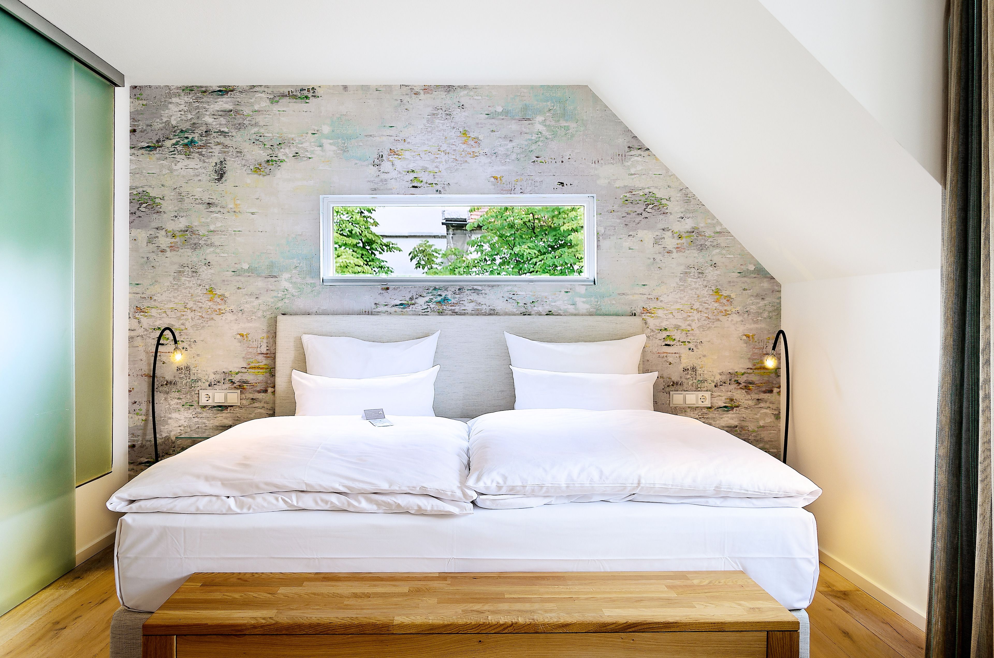 Un dormitorio con cama, un banco de madera, un televisor montado en la pared y un ventanal con vista a árboles.