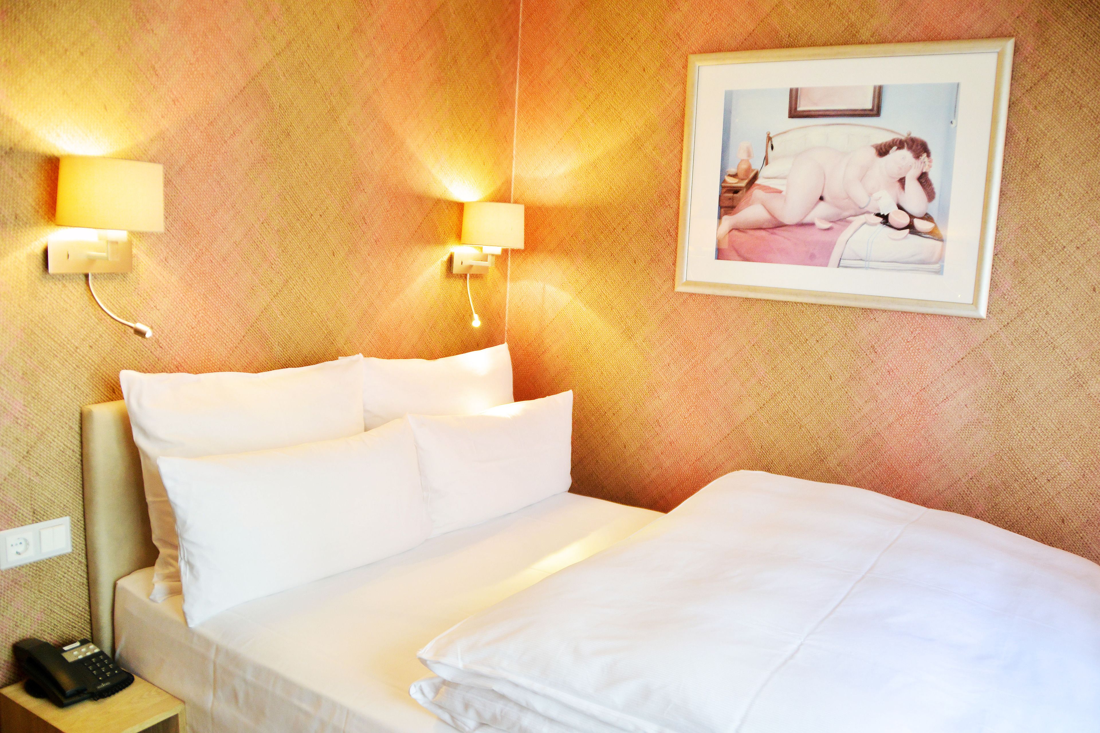 A hotel room with a neatly made bed, two wall-mounted lamps, a framed picture on the wall, and a bedside telephone.