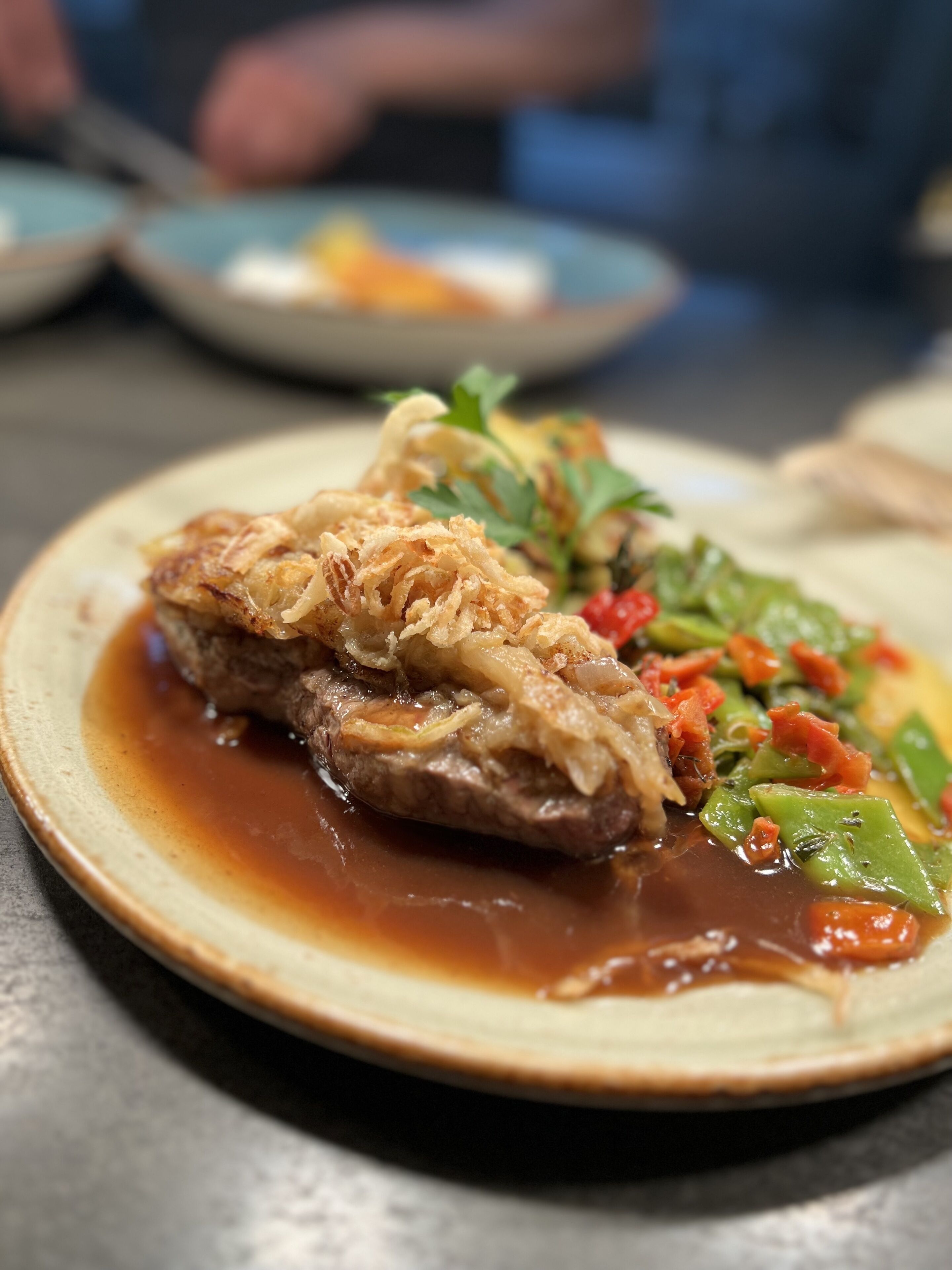 A plate of food with a piece of meat topped with onions, accompanied by a side of vegetables and a sauce.