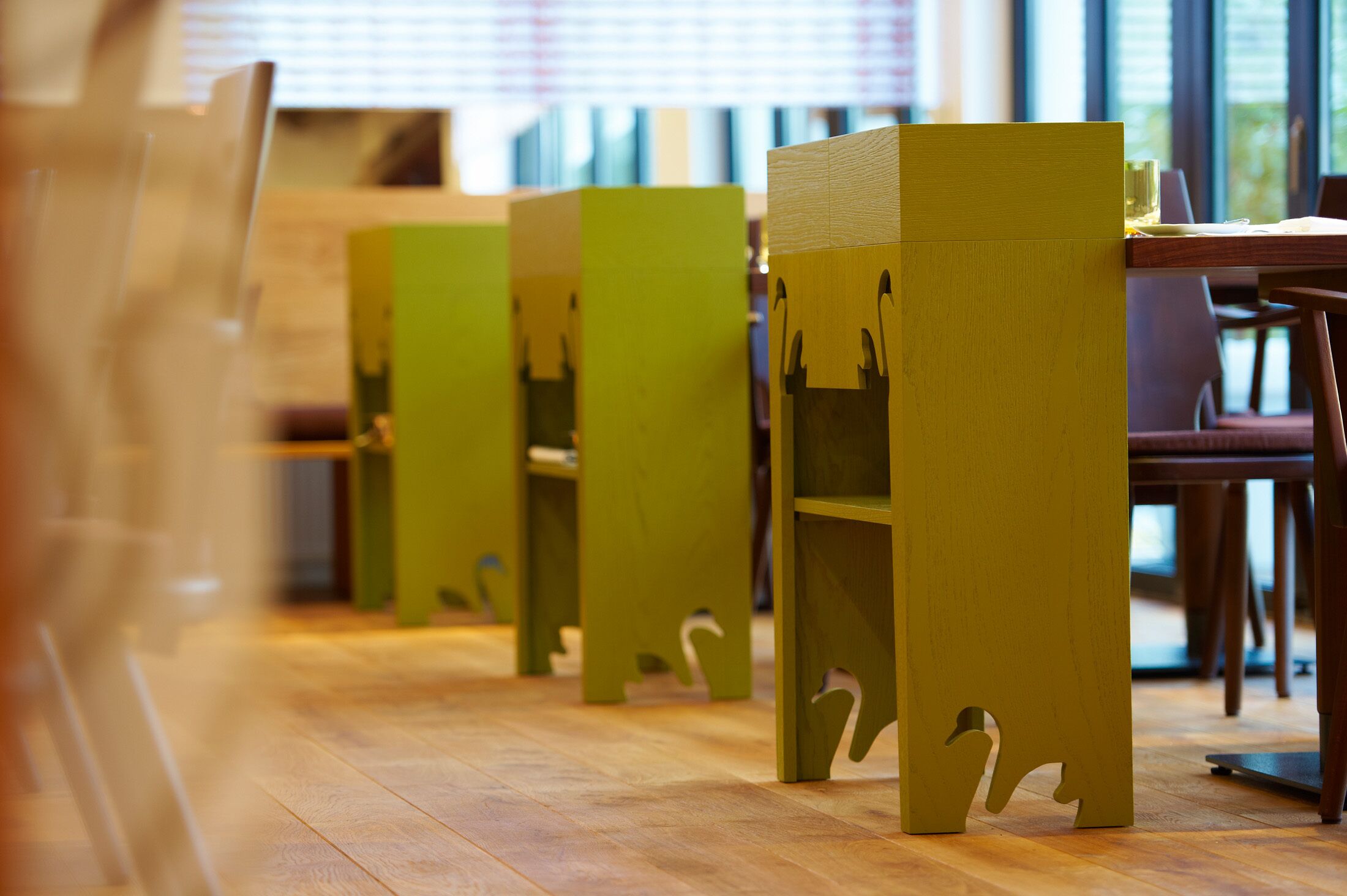 A row of green chairs with cut-out designs on the legs, arranged in a dining area with wooden flooring.