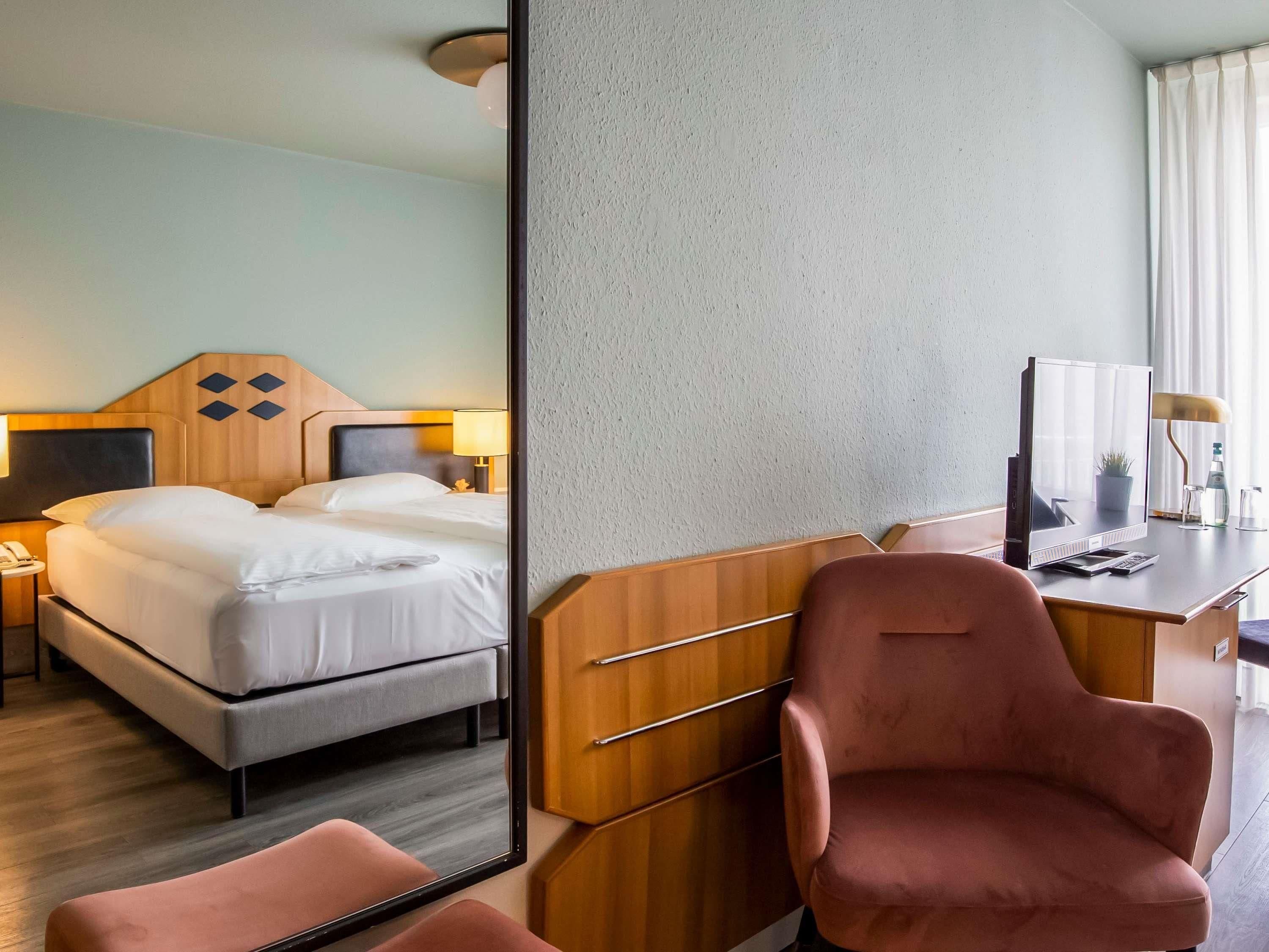 Ett hotellrum med en säng, ett sängbord, en lampa, ett skrivbord, en stol och en stor spegel.