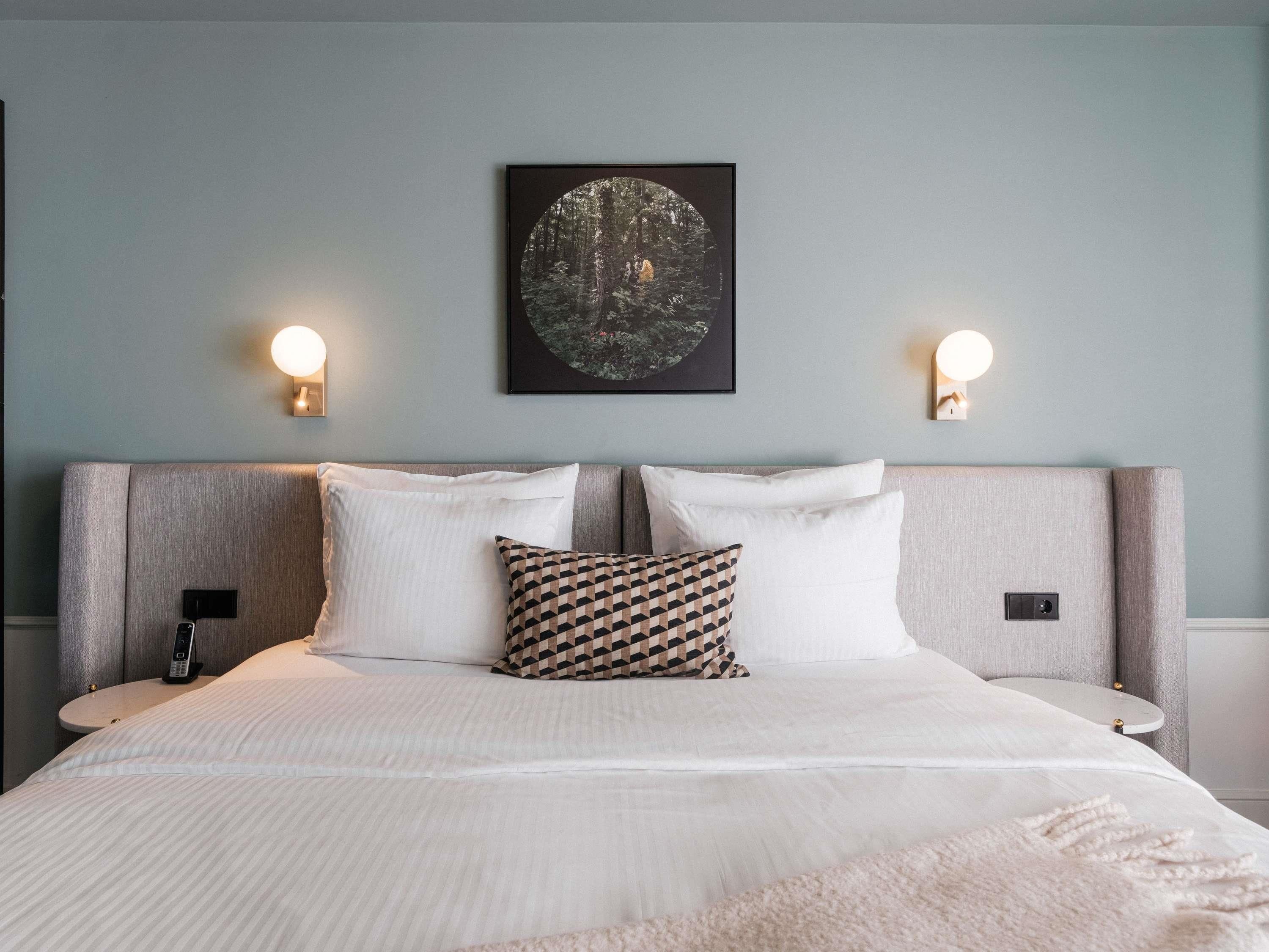 A neatly made bed with white linens and a patterned pillow, a wall-mounted artwork, and two wall sconces.