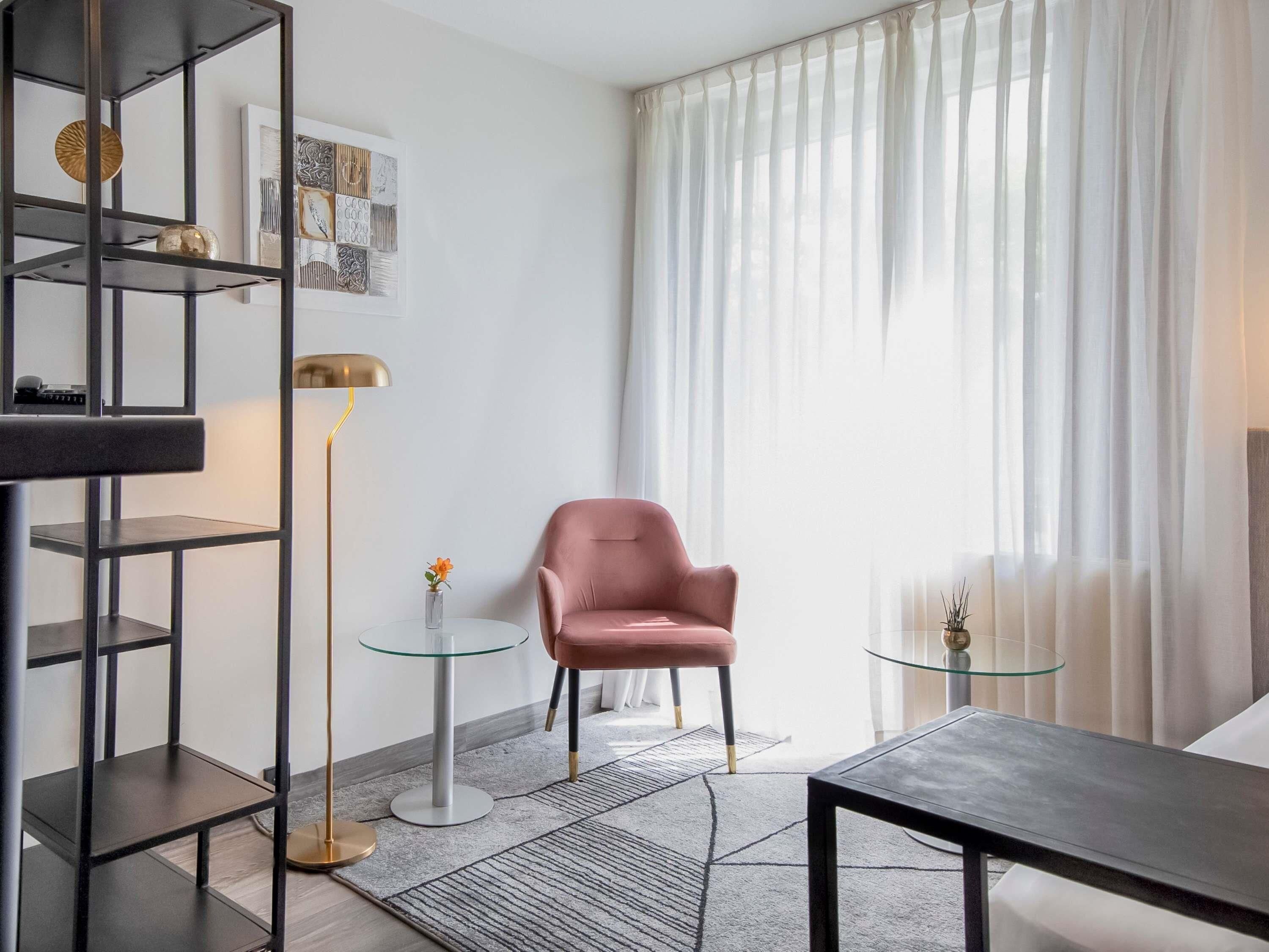 A modern living room with a pink armchair, a glass coffee table, a floor lamp, and a bookshelf.