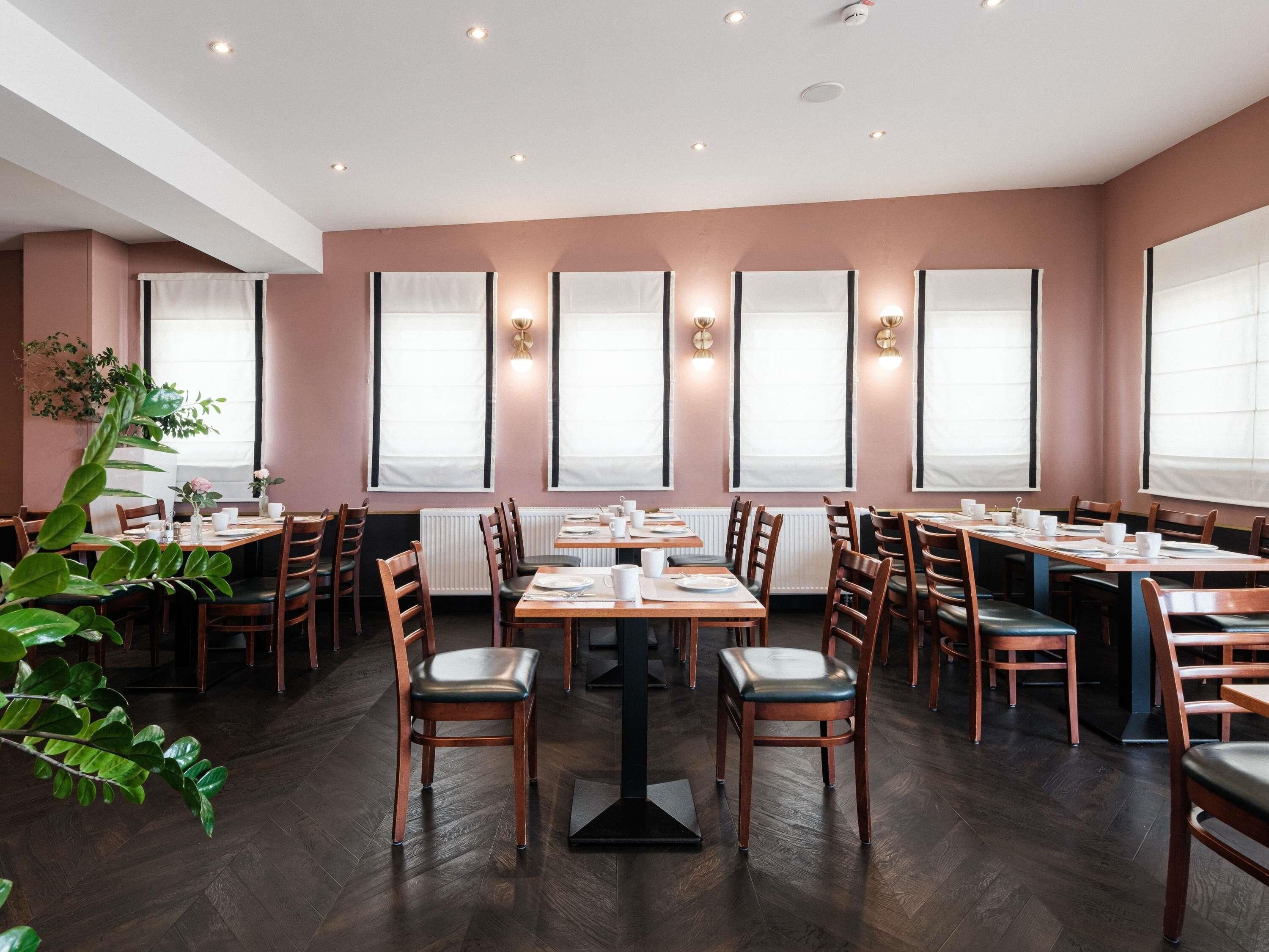 A restaurant interior with wooden tables and chairs, large windows with curtains, and wall-mounted lighting.