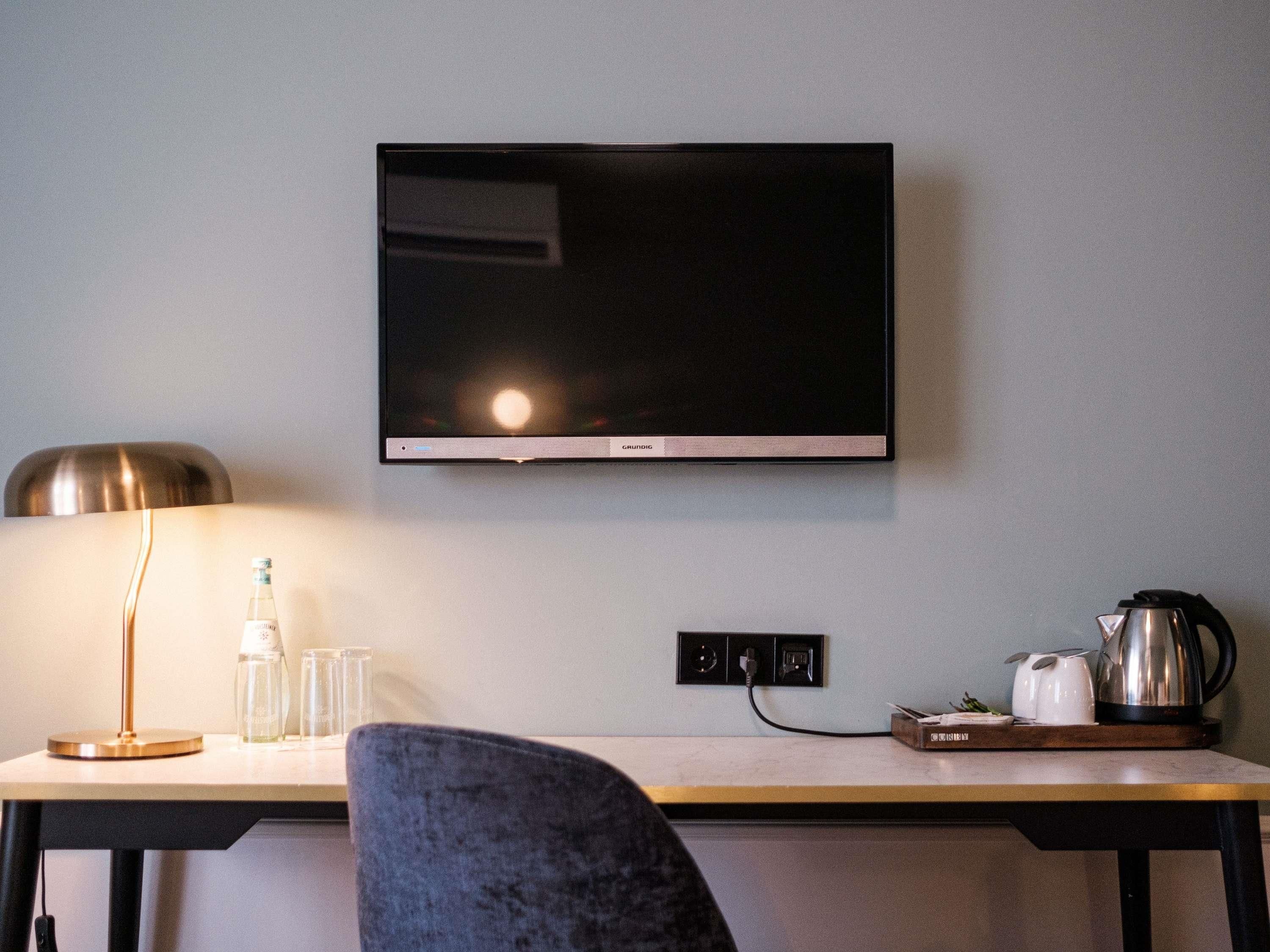 A hotel room with a flat-screen TV mounted on the wall, a desk with a lamp, glasses, and a kettle.