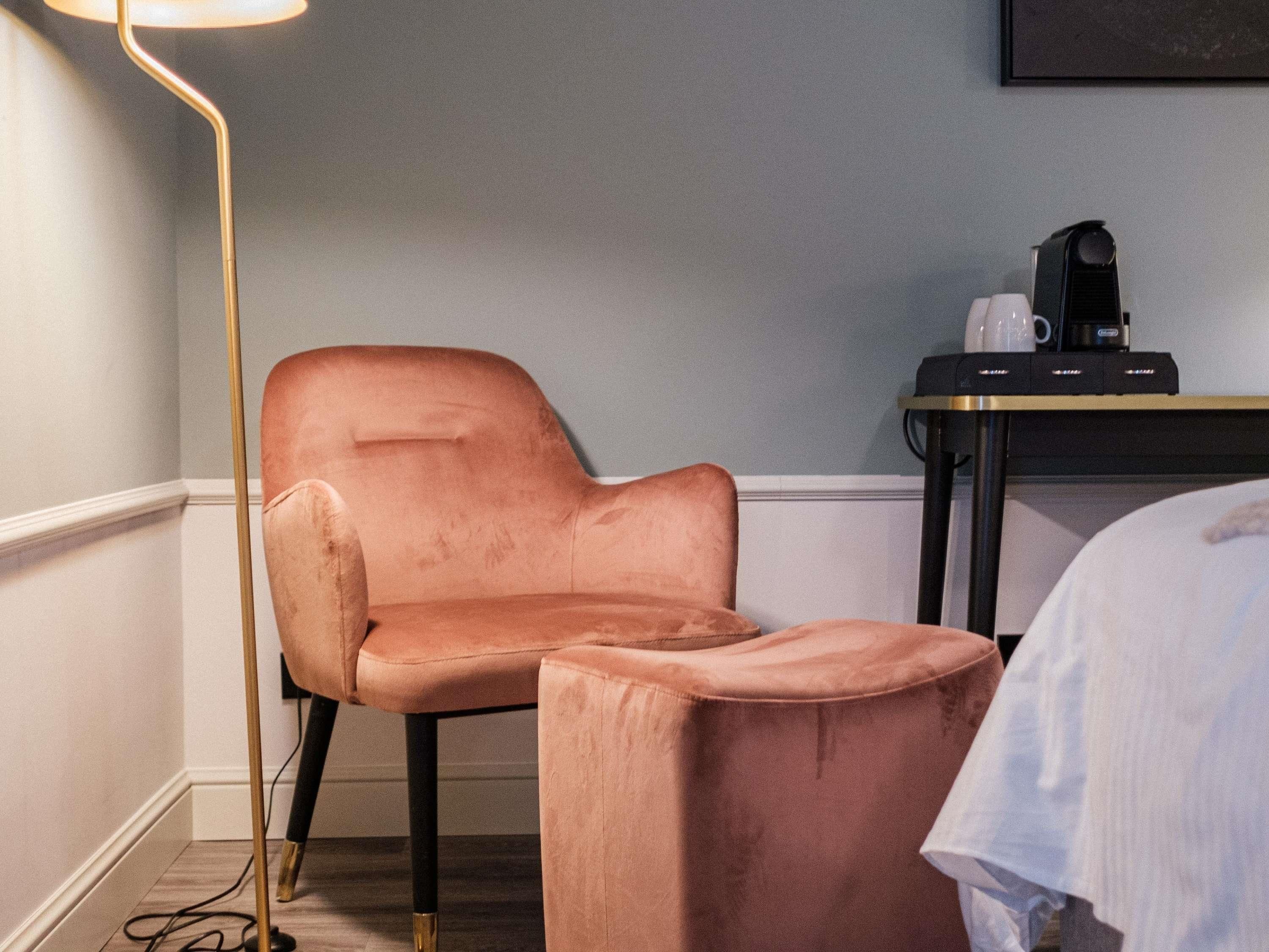 Een kamer met een roze stoel, een klein tafeltje met een koffiezetapparaat en een bed met een witte dekking.
