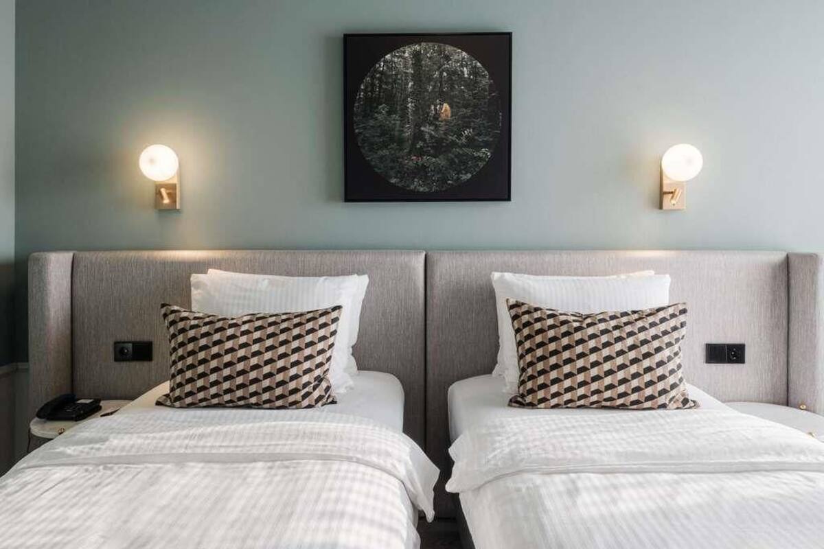 A hotel room with two beds, each with a patterned pillow, a framed artwork above the beds, and wall-mounted lights on either side.