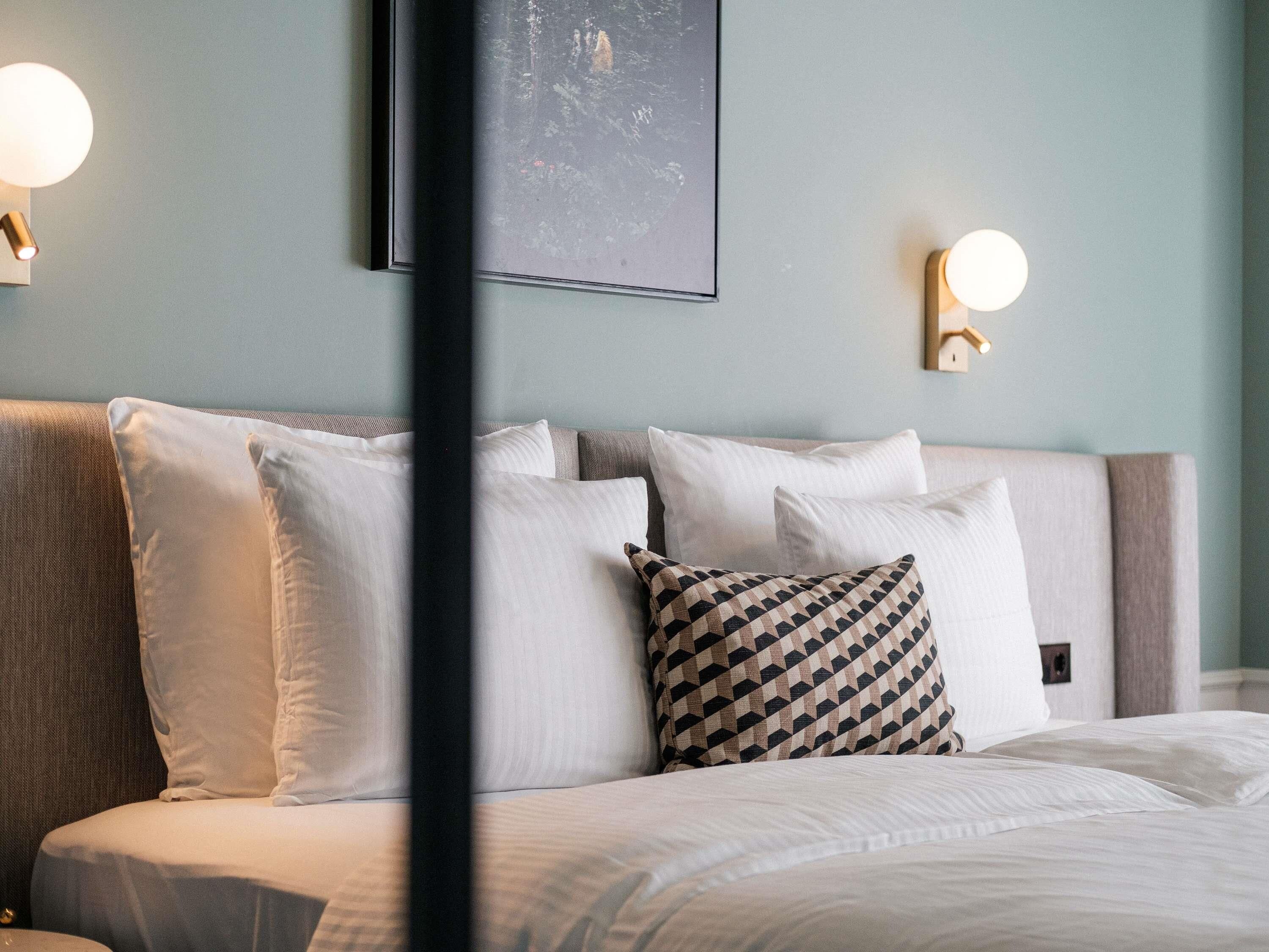 A neatly made bed with white and patterned pillows, a headboard, and wall-mounted lights.
