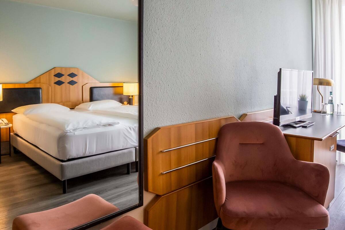 A hotel room with a bed, bedside table, lamp, desk, chair, and a large mirror.