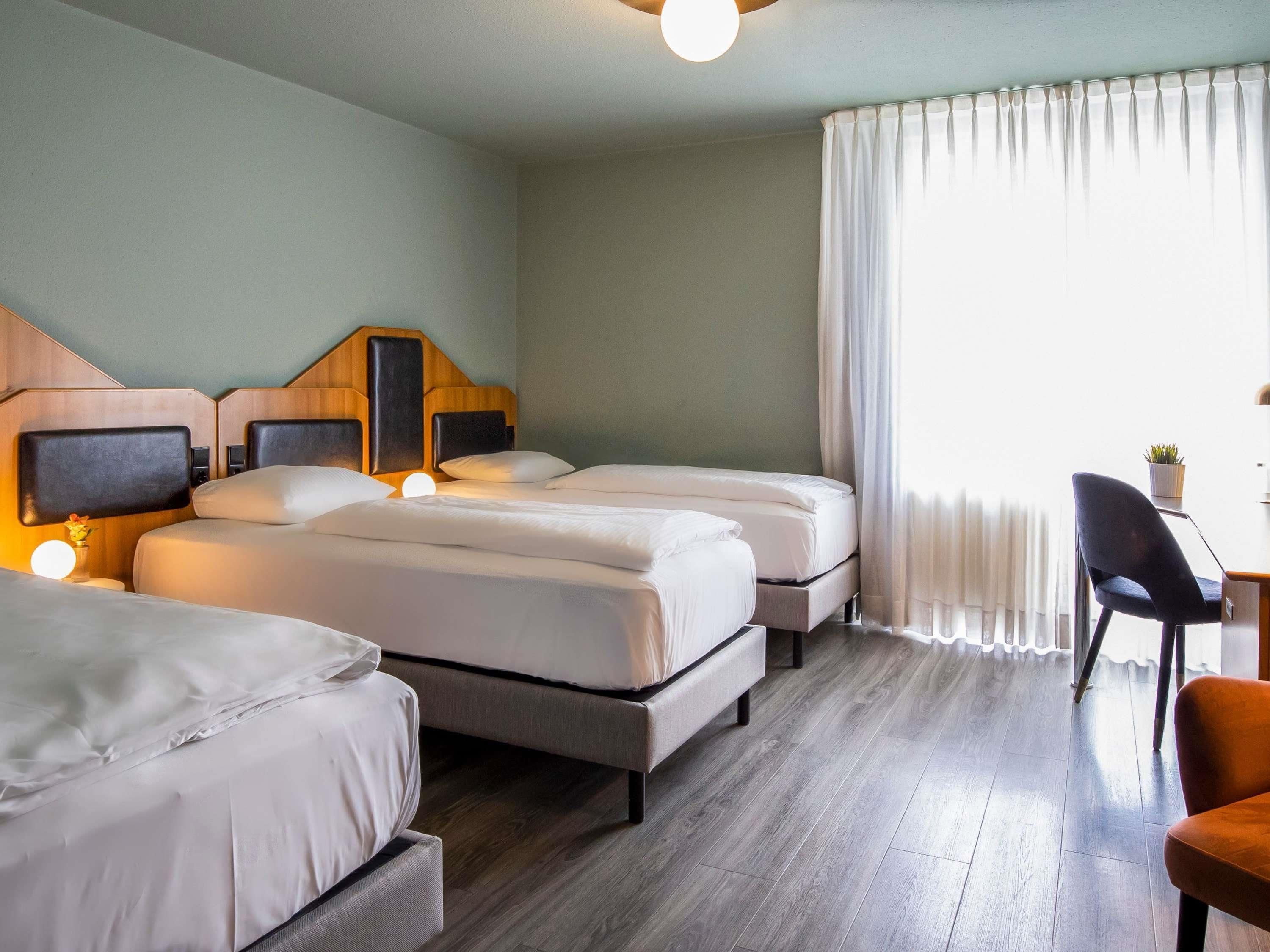 Habitación de hotel con dos camas, un escritorio, una silla y un ventanal con cortinas.
