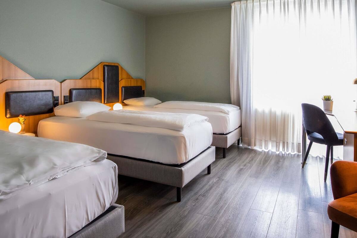 Habitación de hotel con dos camas, un escritorio, una silla y un ventanal con cortinas.