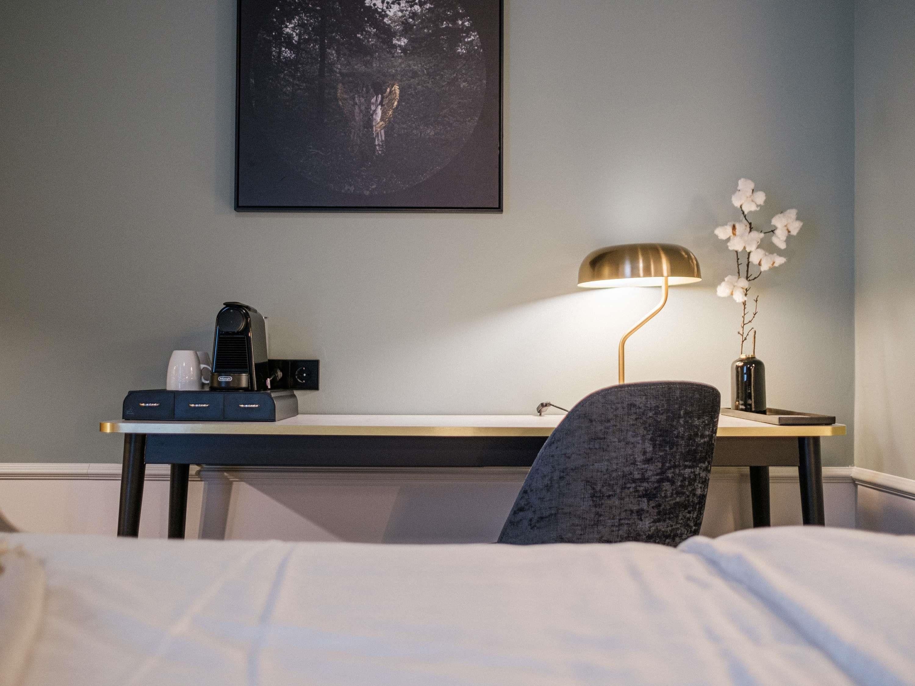 Quarto de hotel com uma cama, uma escrivaninha com luminária, uma cafeteira e um quadro na parede.